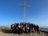 Na szczycie góry stoi duża grupa uczniów liceum. Za nimi metalowa barierka i duży metalowy krzyż. Nad nimi błękitne niebo.
