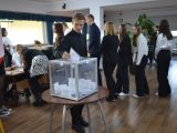Przy stoliku siedzą uczniowie liceum. Przed stolikiem pochyla się dziewczyna i podpisuje kartkę. Chłopak w czarnej koszuli...