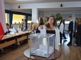 Uczennica liceum wrzuca kartkę do plastkiowej, przezstroczystej urny wyborczej. W oddali stoi grupa osób.
