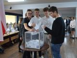 Czterech uczniów liceum wrzuca kartki do plastkiowej, przezstroczystej urny wyborczej. W tle czworo uczniów liceum siedzi...