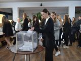 Uczennica liceum w czarnym ubraniu wrzuca kartkę do plastkiowej, przezstroczystej urny wyborczej. W tle w kolejce przed...