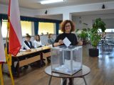 Kobieta w okularach wrzuca kartkę do plastkiowej, przezstroczystej urny wyborczej. W tle flagi: biało-czerwona, żółta.
