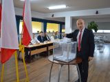Mężczyzna w garniturze wrzuca kartkę do plastkiowej, przezstroczystej urny wyborczej. W tle flagi: biało-czerwona,...