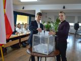 Dwóch uczniów liceum wrzuca kartkę do przezroczystej urny. W tle przy długim stoliku siedzi pięcioro uczniów liceum.