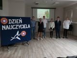 Uczennica w czarnej sukience śpiewa przez mikrofon. Za nią grupa uczniów w strojach galowych. Obok granatowa tablica z...
