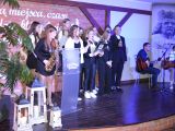 Na scenie stoi grupa uczennic liceum. W grupie stoi dziewczyna i gra na saksofonie. Z prawej stronie przy stoliku siedzi...