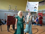 Środkiem sali gimnastycznej idzie dwóch chłopaków. Jeden ubrany w dłuższą kamizelkę, drugi w zieloną sukienkę. Za nimi idą...
