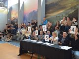 Przy długim stole sidzą dwie kobiety i trzech mężczyzn i uczeń liceum. Za nimi na ławkach siedzą uczniowie liceum.