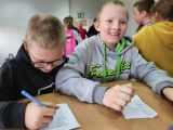 Dwóch uśmiechniętych chłopców z długopisami w dłoniach i białymi kartkami przed sobą siedzi przy stole