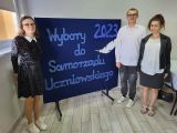 Troje uczniów w strojach galowych. Między nimi granatowa tablica z białym napisem Wybory do Samorządu Uczniowskiego 2023.