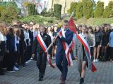Po chodniku uczeń liceum w garniturze i na ramieniu niesie sztandar. Obok niego idą dwie uczennice liceum. W tle duża grupa...