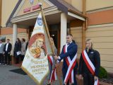 Przed kremowym budynkiem szkoły stoi uczeń liceum w garniturze i trzyma przed sobą sztandar. Obok niego stoją dwie...