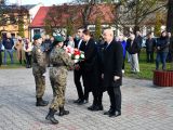 Uczennica liceum w czarnym płaszczu przekazuje biało-czerwone kwiaty trzem osobom w mundurach moro. Obok dziewczyny stoją...