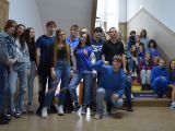 Grupa uczniów liceum stoi na korytarzu szkolnym. Jeden uczeń na pierwszym planie kuca. Wszyscy mają niebieskie ubrania.