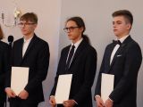 Trzech nastolatków w garniturach stoi w rzędzie na scenie. Przed sobą trzymają dyplomy.