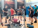 Grupa sportowców w sali gimnastycznej, zgromadzona wokół zawodnika podnoszącego sztange w pozycji leżacej.