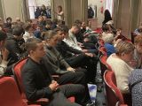 W sali teatralnej w kilku rzędach na czerwonych krzesłach siedzą ludzie.