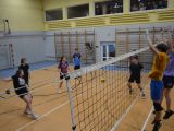 W sali gimnastycznej uczniowie liceum w strojach sportowych grają w siatkówkę. Dwóch chłopców skacze przy siatce.