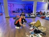 Pośrodku sali mężczyzna w pomarańczowej koszulce kuce i bandażuje nogę dziewczynce siedzącej na prześle. Wokół przy ściaach...