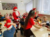 Dzieci siedzą przy stołach. Między nimi Mikołaj i dziewczyna z koszykiem.