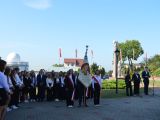 Na dziedzińcu szkoły stoi poczet sztandarowy. W tle grupa uczniów liceum ubranych w czarno białe stroje.