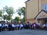 Na dziedzińcu stoi bardzo duża grupa uczniów liceum. Przed uczniami idzie poczet sztandarowy. Z prawej strony budynek szkoły.
