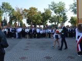 Na dziedzińcu stoi bardzo duża grupa uczniów liceum. Przed uczniami idzie uczeń z kwiatami. Z prawej strony budynek szkoły.