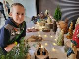 Uśmiechnięty chłopiec oparty łokciami o długi stół przykryty białym obrusem. Na stole bożonarodzeniowe ozdoby wykonane z...