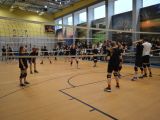 W sali gimnastycznej uczniowie liceum w strojach sportowych grają w siatkówkę. Na trybunach siedzą uczniowie liceum.