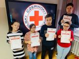 Uczniowie szkoły podstawowej z kolorowymi dyplomami w dłoniach. W tle logo Polski Czerwony Krzyż