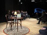Na pierwszym planie stylowy okrągły stół i krzesło. W tle kilkanaście osób, siedzących na krzesłach.