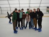 Grupa osób stoi pośrodku lodowiska. Wszyscy mają łyżwy na nogach.