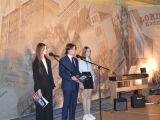Przy dwóch mikrofonach stoi uczeń liceum w garniturze i dwie uczennice liceum, jedna w białej bluzce, druga w czarnej...