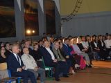 Uczniowie i nauczyciele liceum siedzą na krzesiałach w rzędach w sali gimnastycznej.