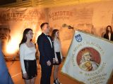 Uczeń w garniturze i dwie uczennice w białych bluzkach stoją przed opuszczonym sztandarem.