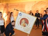 Uczeń w garniturze trzyma opuszczony sztandar, drugi uczeń całuje sztandar. Obok niego stoją dwie uczennice w białych...