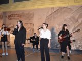 Pośrodku sali dwie stoją dwie dziewczyny i śpiewają do mikrofonów. Z tyłu dwie dzewczyny grają na gitarach, a jedna na...