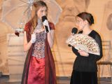 Pośrodku sali stoją dwie licealistki w strojach dworek. Jedna trzma letni parasol, druga wachlarz.