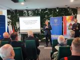 Sala konferencyjna. Zdjęcie z tyłu. 2 prowadzących zwróconych twarzami do słuchaczy.