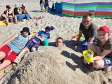 Grupa uczniów siedzi na plaży letnich strojach. W tle parawany