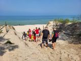 Grupa uczniów z plecakami i czapkami na głowach schodzi piaszczystym zejściem na plażę.W tle morze