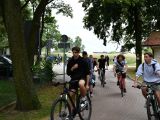 Uczniowie liceum, szkoły podstawowej i dorośli jadą na rowerach aswaltową drogą. Po obu stronach drogi drzewa.