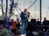 Pośrodku sceny stoi mężczyzna w niebieskiej marynarce i mówi do mikrofonu. W tle orkiestra.