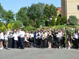 Na dziedzińcu szkoły stoi duża grupa uczniów liceum w strojach szkolnych (chłopcy w garniturach, dziewczyny w białych...
