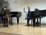 Mężczyzna w jasnej marynarce siedzący przy fortepianie. Obok kobieta ze skrzypcami w dloniach